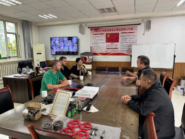 Five people are sitting around a table in an office. Three men and two women are engaged in a discussion. On the table are various tools and documents, as well as several cups of drink. On the wall is a large screen showing a surveillance image and an information poster in red and yellow related to an organization. The room has windows that let in natural light.