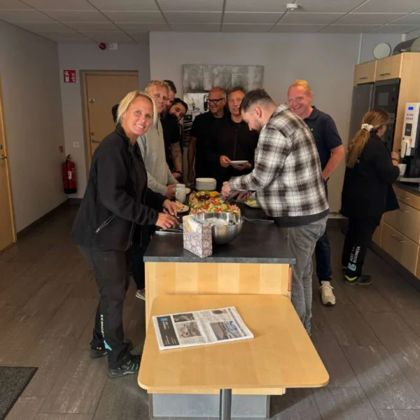 A group of people gather around a dining table in a kitchen. Several people smile and serve themselves from a large salad bowl. The table is covered with plates and a newspaper. A smoke alarm is visible on the wall in the background, and a woman stands by a coffee machine. The room has a neutral color scheme and modern kitchen furniture.