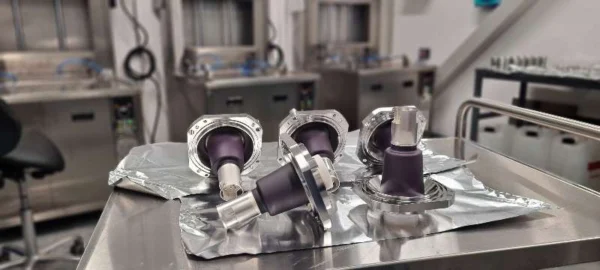 Flera svarta och silverfärgade delar med runda fästen ligger på ett metallbord täckt av aluminiumfolie. I bakgrunden kan man se laboratorieutrustning och arbetsstolar i en renrumsmiljö.