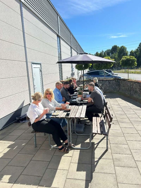 A group of people are sitting at a table outside, under a parasol, on a sunny day. They are eating and drinking coffee, and there are several plates of food in front of them. The background shows a gray building with large windows and a stone wall, as well as some trees and a car in the parking lot. Spring or summer clothes are visible on the participants.