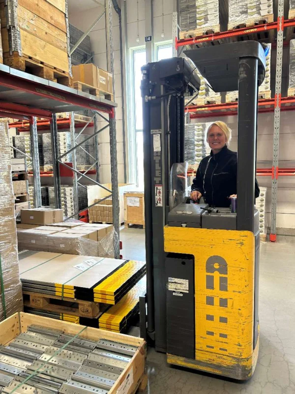 En kvinna med blont hår står vid en gaffeltruck i ett lager, omgiven av staplar med varor och pallager. I bakgrunden syns hyllor fyllda med lådor och produkter, medan några pallar med förpackningar är placerade på golvet. Belysningen i rummet är naturlig från fönsterna.