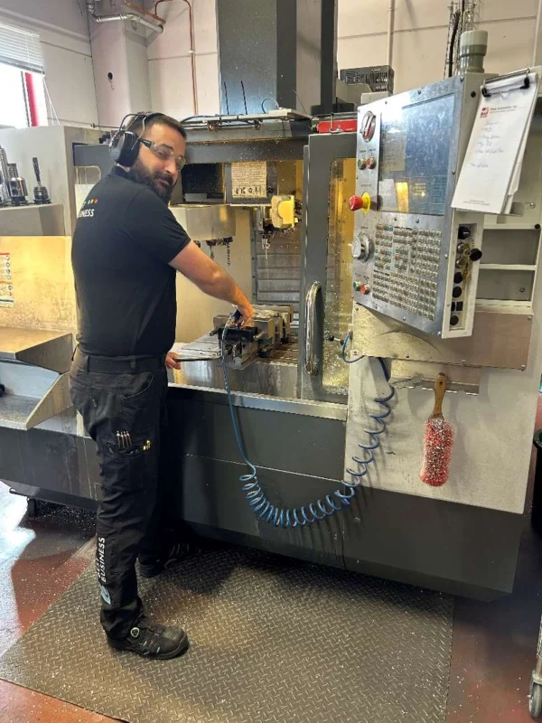 A man with a beard and ear protection is standing at a CNC machine working on a workpiece. He is wearing a black t-shirt and work pants with tools in his pockets. The machine is gray and has a digital control panel with several buttons and screens. There are chips and liquid on the work surface, and in the background there are tools on shelves. The room appears to be a workshop with an industrial interior.