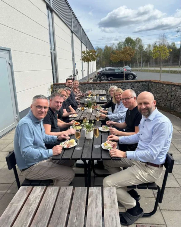 Ett gäng personer sitter vid ett långbord utomhus på en solig dag, omgivna av en grönskande miljö. De äter middag och har tallrikar med mat framför sig, samt glas med dryck. Det finns blommor i en kruka på bordet. I bakgrunden syns en byggnad och en parkerad bil. Runt bordet finns både män och kvinnor, som verkar ha en trevlig stund tillsammans.