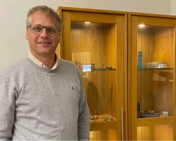 A man with glasses and a gray shirt stands smiling in front of two wooden cabinets filled with metal objects and mechanical parts.