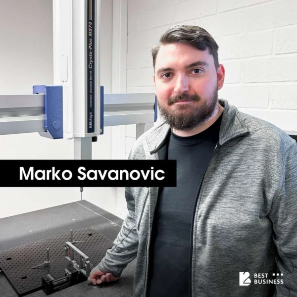 A man with short, dark hair and a beard stands by a measuring machine with a large platform and tools. He is wearing a gray jacket and looking at the camera. In the background is a white brick wall.