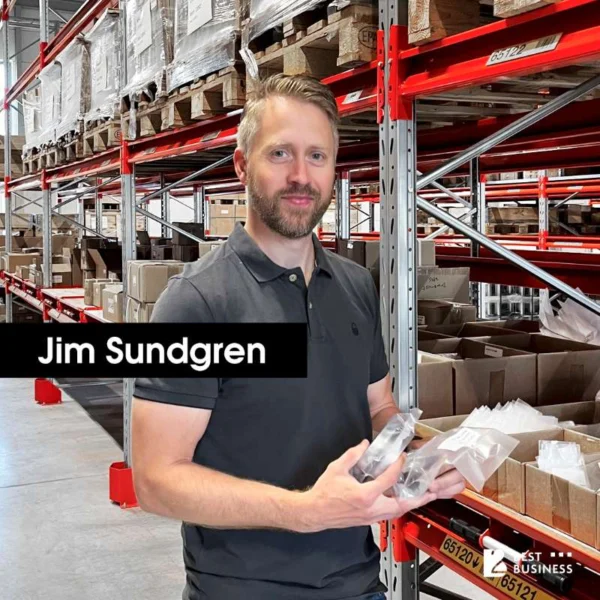 En man med kort hår och skägg står i en lagerlokal. Han håller i några plastförpackningar och ser mot kameran med ett leende. I bakgrunden syns hyllor med lådor och pallar, samt ett ordnat lagerutrymme. En markerad text i bildens nedre del visar namnet "Jim Sundgren".
