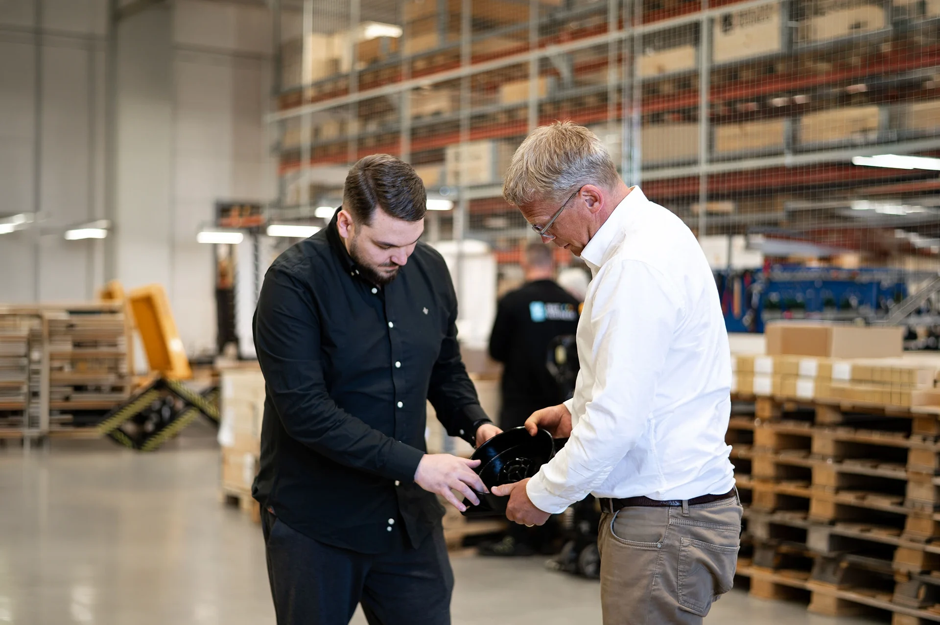 Två män står i en lagerlokal, båda fokuserade på en svinande svart komponent som de håller i händerna. Den ena mannen har på sig en svart skjorta och den andra en vit skjorta. I bakgrunden syns staplade pallkedjor och hyllor med lådor, vilket ger en känsla av en aktiv arbetsmiljö.